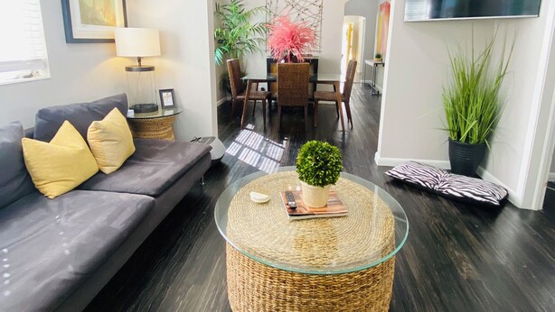 Una sala de estar moderna con piso de madera oscura, una mesa de centro de vidrio, un sofá gris con cojines amarillos, una zona de comedor con candelabro y un televisor de pantalla plana montado en la pared.