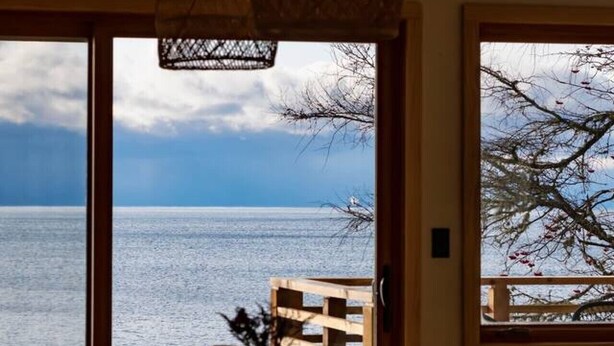 Un coin repas avec une table et des chaises en bois, un vase rempli de fleurs et une vue sur la mer grâce à de grandes fenêtres.