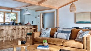 Un intérieur moderne avec un plafond en bois, un coin cuisine doté de tabourets de bar, un canapé avec des coussins à motifs et un luminaire suspendu.