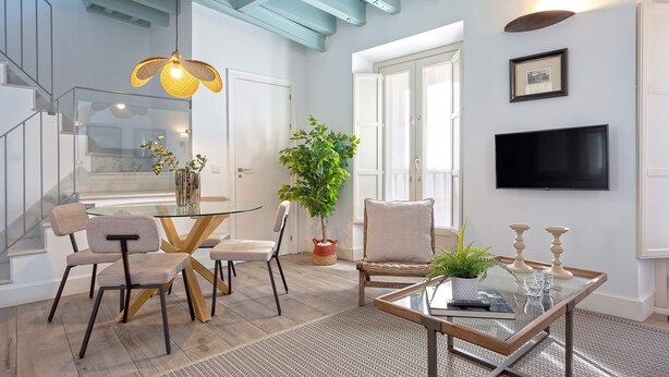 Una sala de estar moderna con una mesa de centro de vidrio, un comedor con una mesa y sillas de vidrio, una planta grande en maceta y un televisor de pantalla plana montado en la pared.