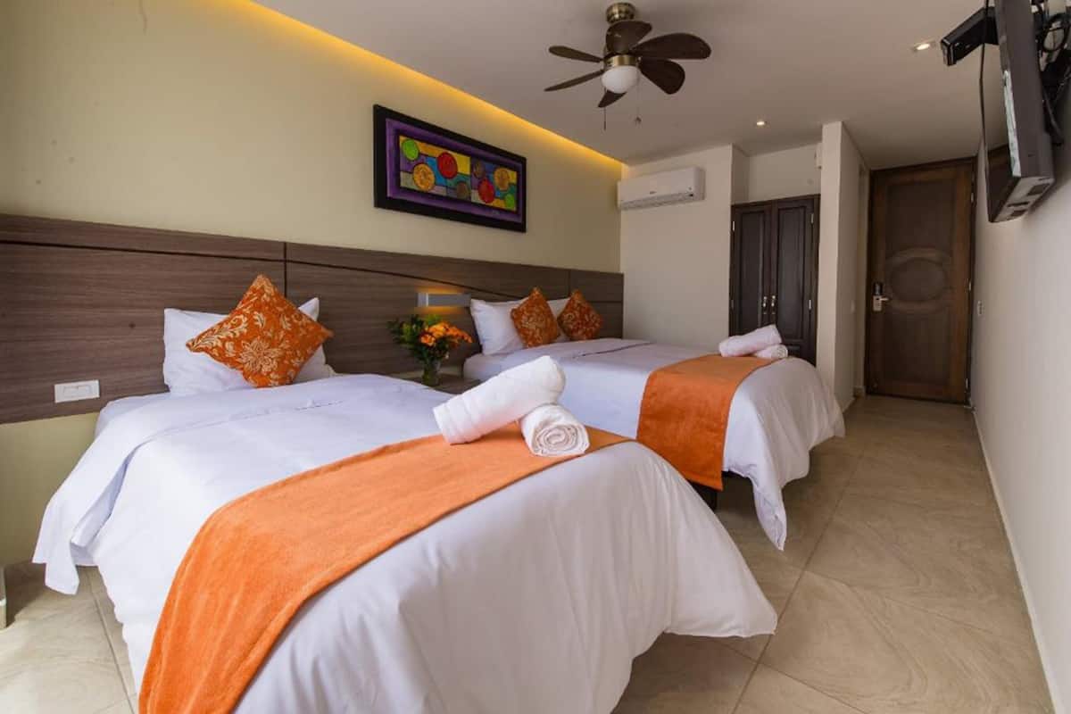A hotel room with two beds, each with white bedding and orange throw blankets, a ceiling fan, a wall-mounted TV, and a framed artwork above the beds.