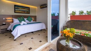 A hotel room with a bed, a nightstand, a wall-mounted air conditioner, a sliding glass door to an outdoor area, and a wicker chair with a table and vase of flowers.
