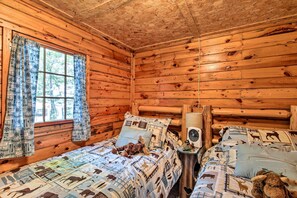 A cozy cabin room with wooden walls, a bed with patterned bedding, a small table with a fan, and a window with curtains.