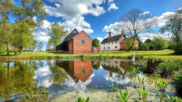 A serene lakeside setting with a brick barn and a white house, surrounded by lush greenery and a clear sky. - 16th century grade II* farmhouse with hot tub and wild swimming (Harleston)