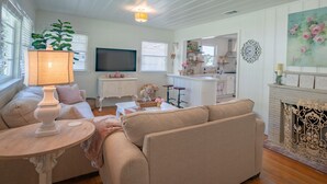 Ein Wohnzimmer mit einer Couch, einem Tisch mit einer Lampe, einem Fernseher und einem durchgegangenen Tür erkennbaren Küchenbereich.