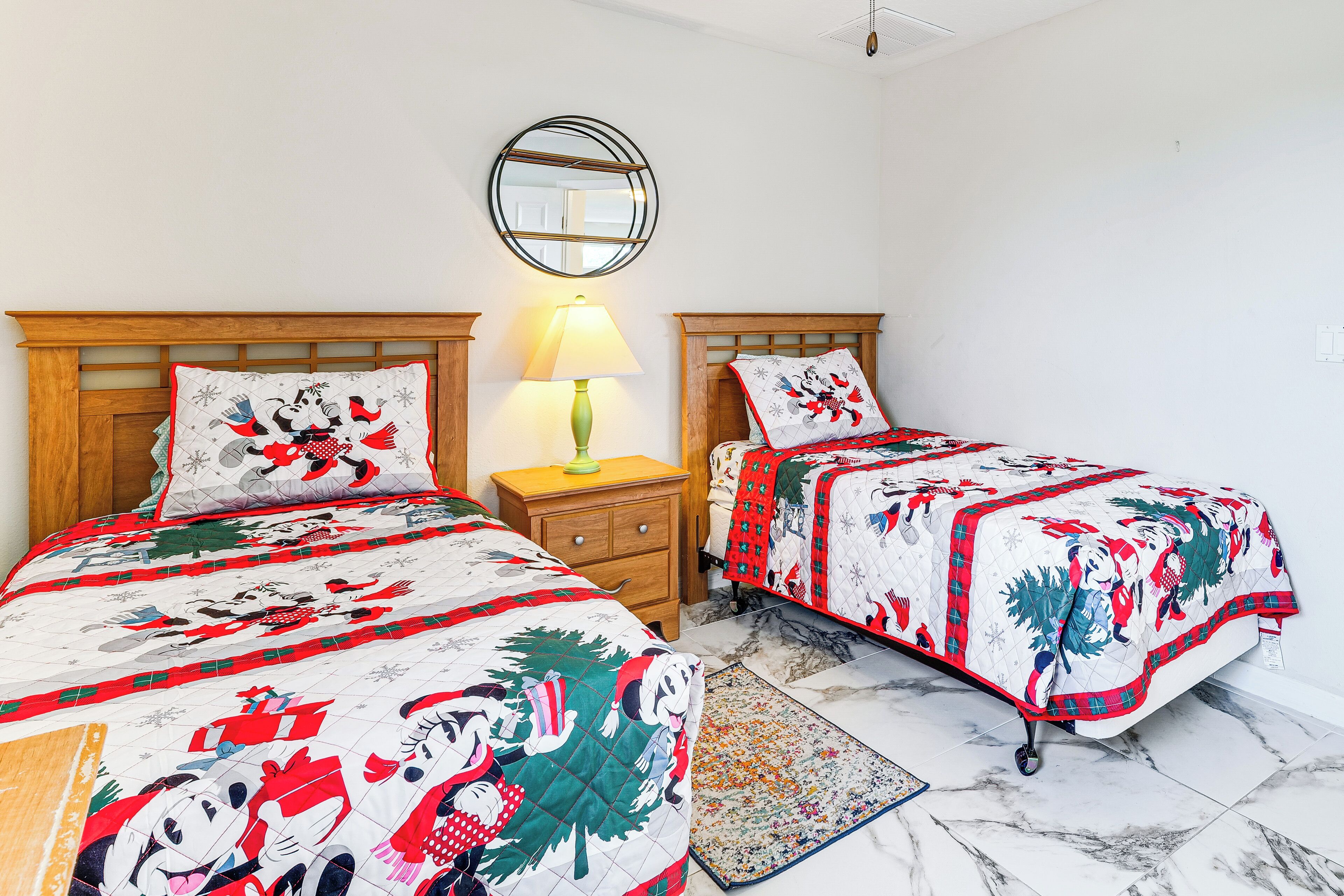 Two beds with matching quilts, a bedside table with a lamp, a round mirror on the wall, and a patterned rug on the floor.