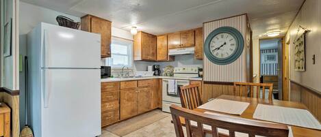 A compact kitchen with wooden cabinets, a white refrigerator, a dining table with chairs, and a wall clock.