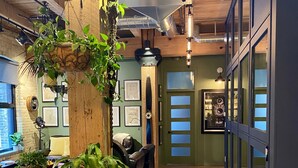 A modern interior with wooden floors, a central wooden column with hanging plants, a seating area with a sofa and armchairs, and a view through a glass door to a room with a large mirror and decorative items. - theLOFTonJAMES, Historic Exchange District, Exposed Brick & Beams, High Ceilings (Winnipeg)