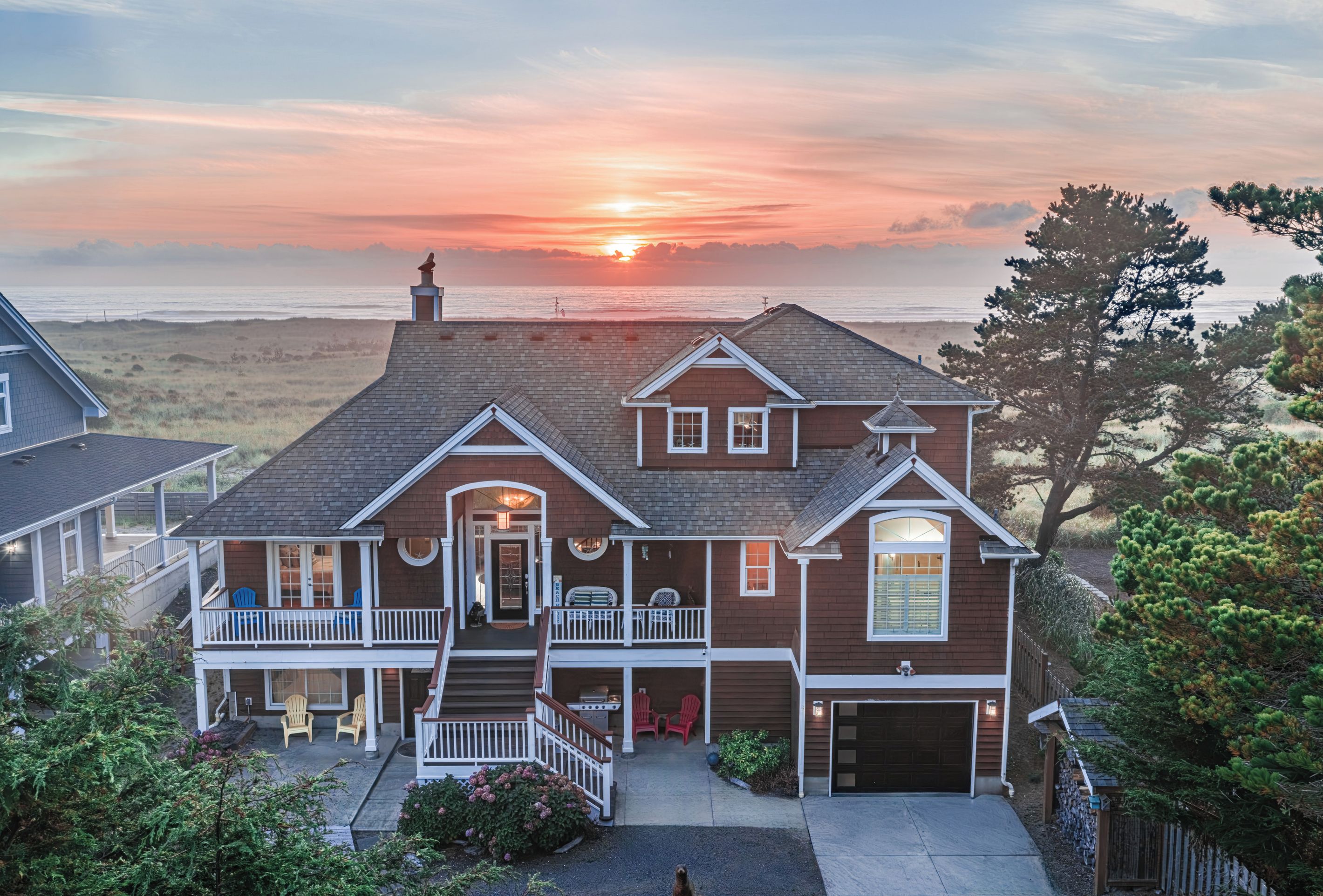 Une maison à deux étages avec un porche à l’avant, un garage et une vue sur l’océan au coucher du soleil.