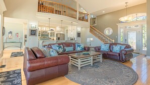 A spacious living area with a large brown leather sofa, a coffee table, and a patterned rug. A kitchen with white cabinets and a dining area with a glass table are visible. A staircase leads to an upper level with a wooden railing. - Coral Sunsets & Shimmering Deep Blue Ocean Views! (Ocean Park)