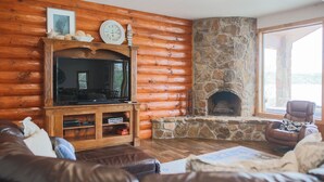 Interior de una cabaña de madera con chimenea de piedra, paredes de madera, un sofá de cuero y un televisor sobre un mueble de madera.