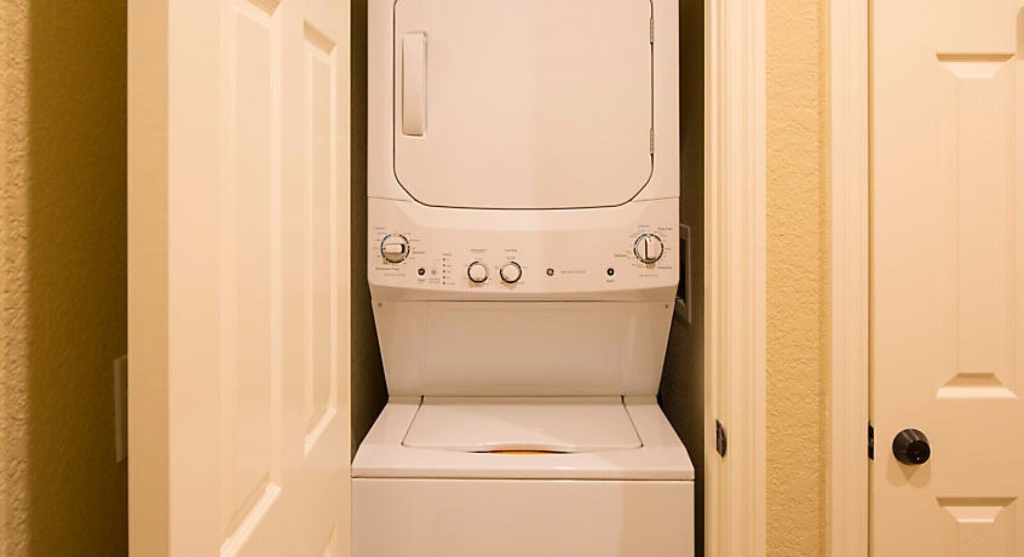 A compact, white, front-loading washer and dryer unit in a narrow hallway with beige walls and a door on the right.
