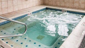 Un pequeño jacuzzi interior con una escalera de metal, paredes de azulejos y una ventana con vistas a edificios.