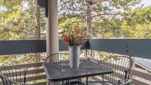 Un espacio de estar al aire libre cubierto con una mesa y sillas de metal, un cubo de metal con flores rojas, y una vista de árboles y un edificio al fondo.