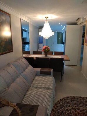 A modern living room with a sofa, a dining table with chairs, a chandelier, and a kitchen in the background. - High level apartment, air cond. in every room and sea view (Bombinhas)