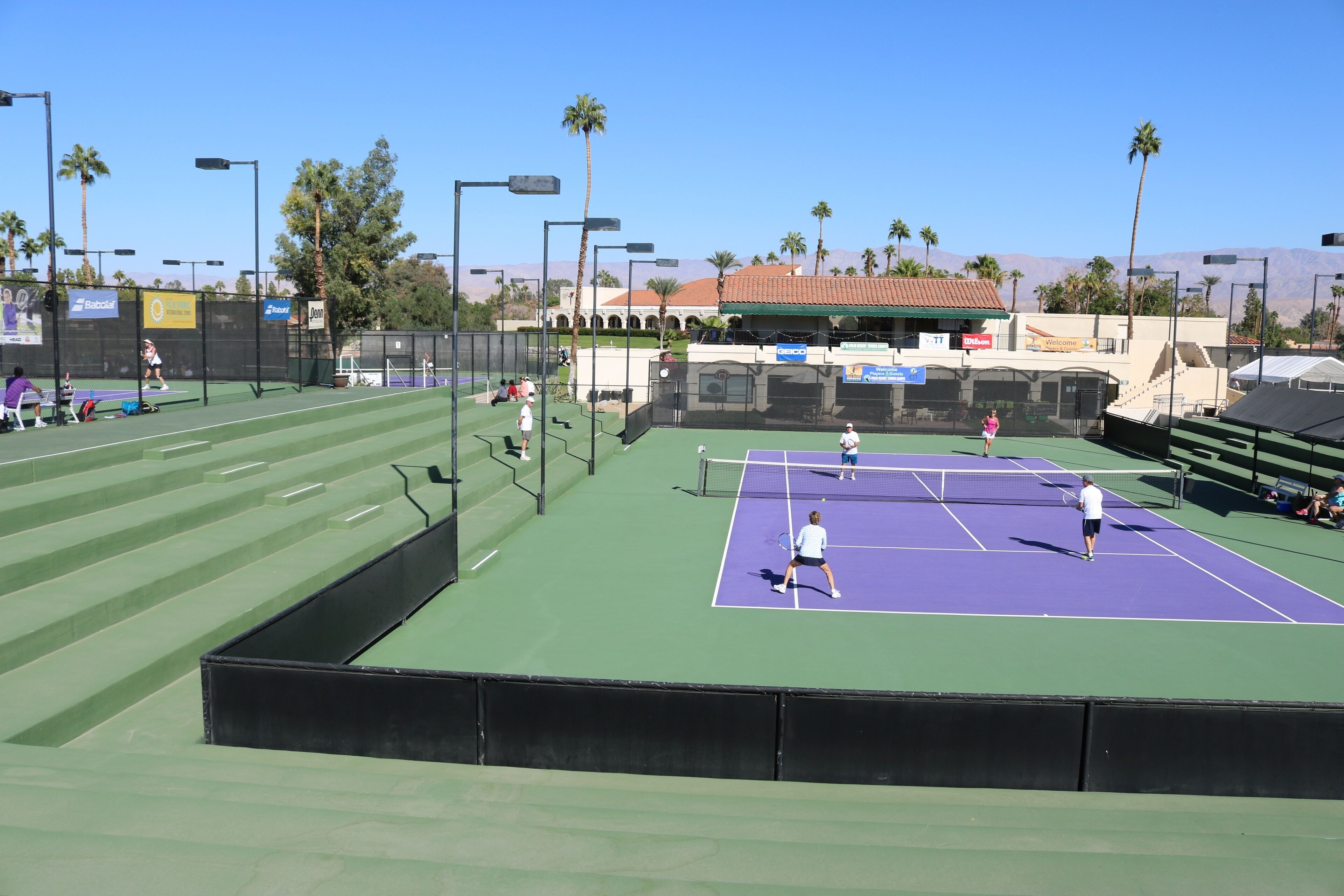 Ein Tennisplatz mit Netz, Sitzbereichen und Palmen im Hintergrund.