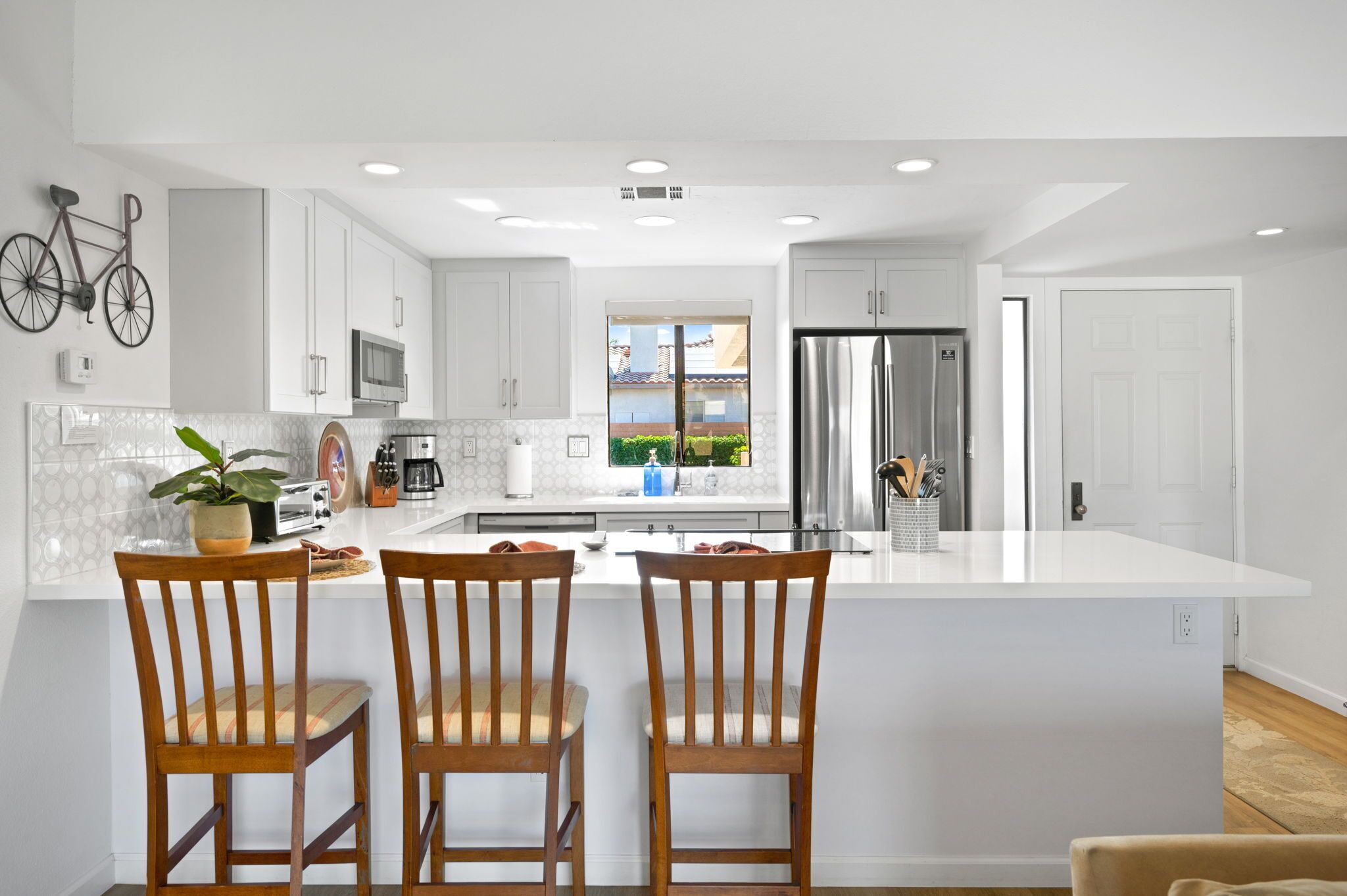 A modern kitchen with white cabinetry, a large island with bar stools, a microwave, a refrigerator, and a window with a view of a house and greenery.