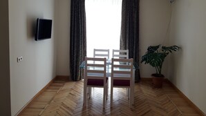 A room with a herringbone wooden floor, a dining table with two chairs, a potted plant, a wall-mounted TV, and a window with patterned curtains. - Cozy clean and bright studio8 (Arabkir)