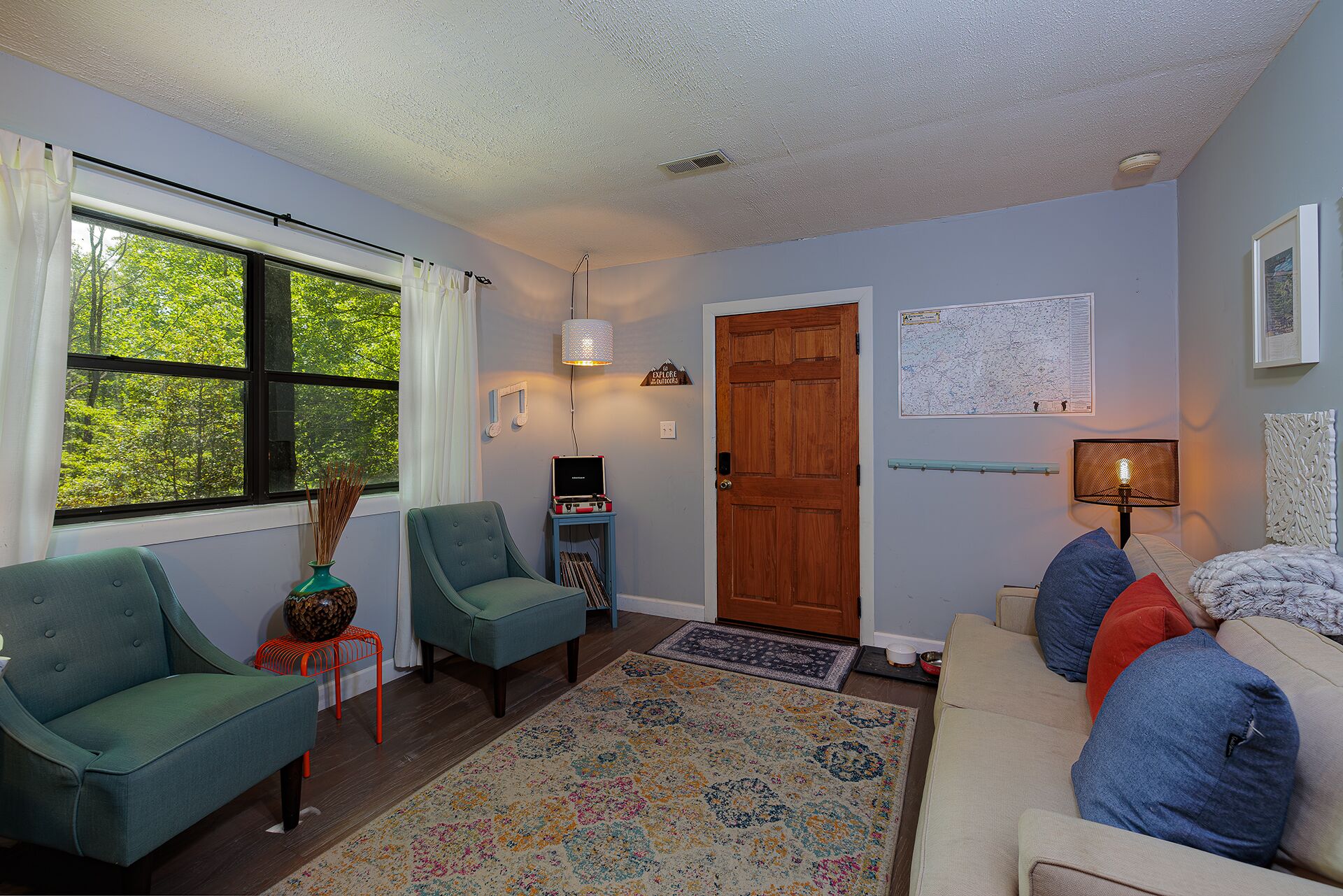 A living room with a sofa, chairs, a rug, a wooden door, a map on the wall, and a window with a view of trees.