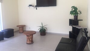 Un soggiorno con una TV a schermo piatto montata a parete, due sgabelli di legno, un divano nero, una pianta in vaso e un piccolo ripiano con oggetti.