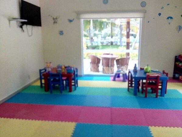 Ein Zimmer mit einem bunten Spielbereich, zwei Tischen mit Stühlen und einem großen Fenster mit Blick ins Freie.
