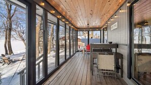 Un espacio exterior cubierto con suelo de madera, ventanas amplias y una zona de estar con mesas y sillas.