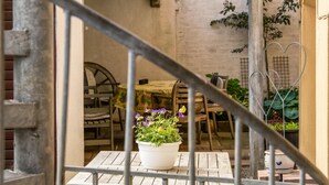 Un espacio de estar rústico al aire libre con una mesa y sillas de madera, una planta en maceta con flores moradas y una baranda de metal.