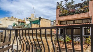 A balcony with a wooden chair and a cushioned bench, overlooking a cityscape with buildings and a clear sky. - Perfectly Located Sliema Apartment (Sliema)