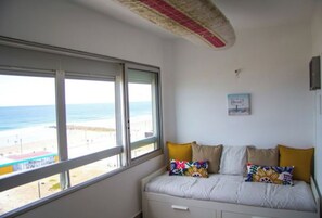 A room with a large window overlooking a beach, a bed with colorful pillows, and a small wall-mounted picture.