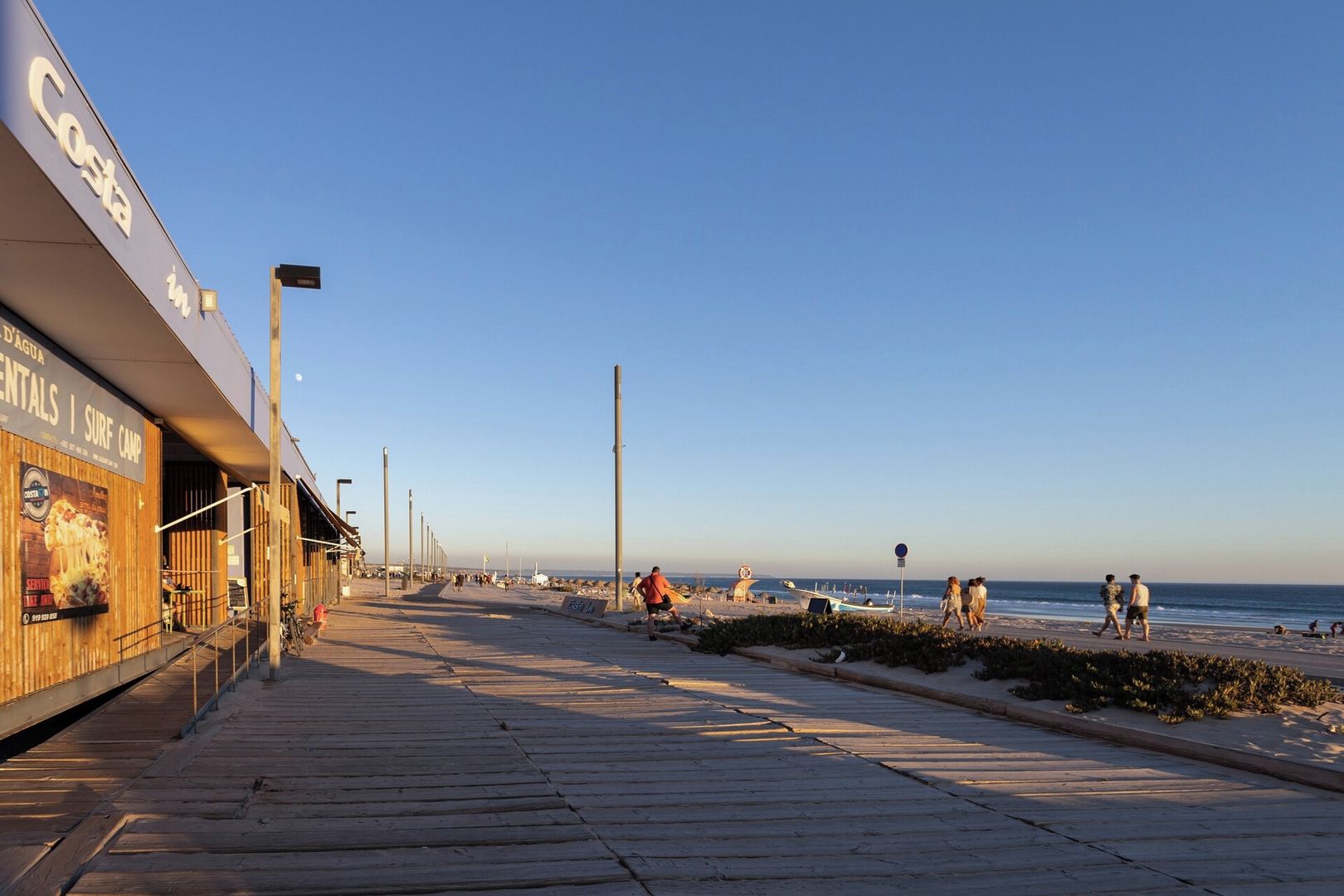 A coastal walkway with a beach on one side, a boardwalk, and a building with a sign for Costa in the background.
