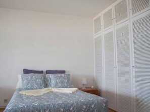 Quarto com cama, duas almofadas, mesa de cabeceira com abajur e armário branco com portas venezianas.