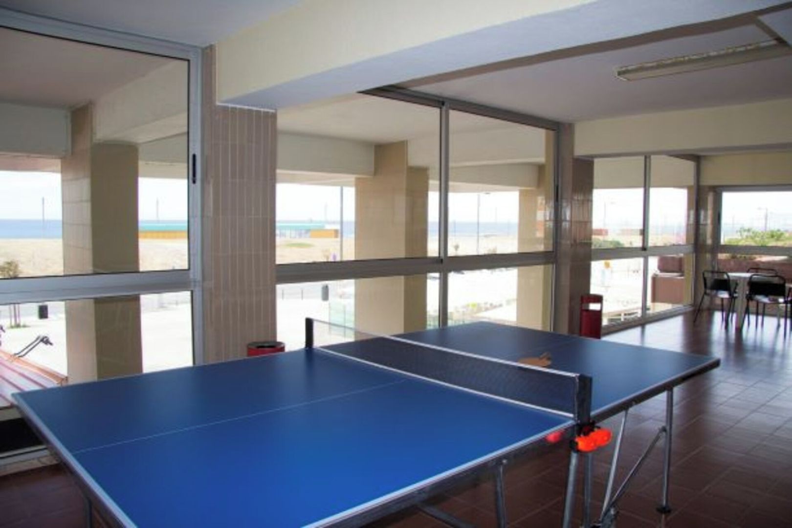 A room with a ping pong table, large windows, and a view of the beach.