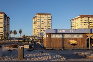 Um imóvel à beira-mar com uma passarela de madeira que leva a um edifício chamado 'Costa', oferecendo aulas de surf, aluguel de equipamentos e um acampamento de surf.