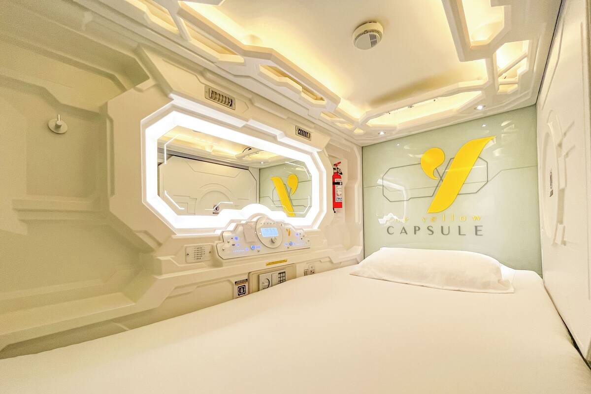 A capsule hotel room with a bed, a mirror, a control panel, and a wall with a yellow logo.