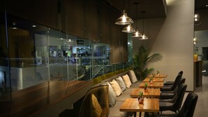 Un restaurante moderno con una larga barra, varias mesas y sillas. Hay lámparas colgantes y un gran ventanal con vistas a una calle concurrida.