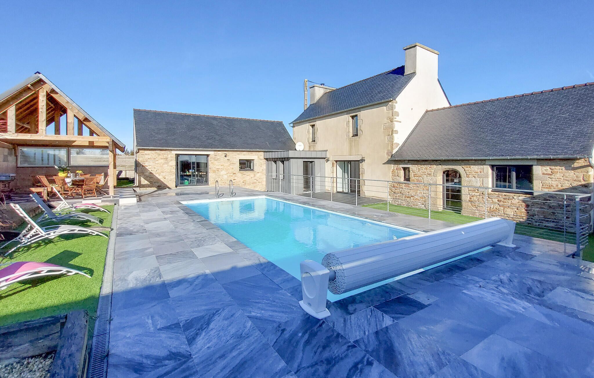Ein Pool mit einer Steinterrasse, ein Holzdeck mit Liegestühlen und ein Steinhaus mit grauem Dach.
