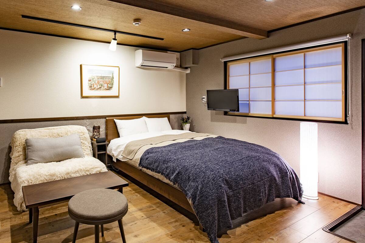 A compact hotel room with a bed, a small seating area, a bedside table, an air conditioning unit, a television, and a window with a view.