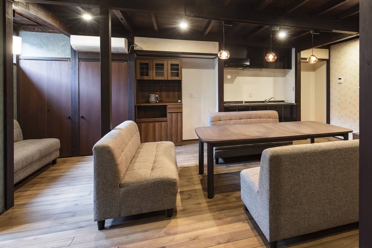 A room with a wooden floor, two sofas, a dining table, and a kitchen area with cabinets and a stove.