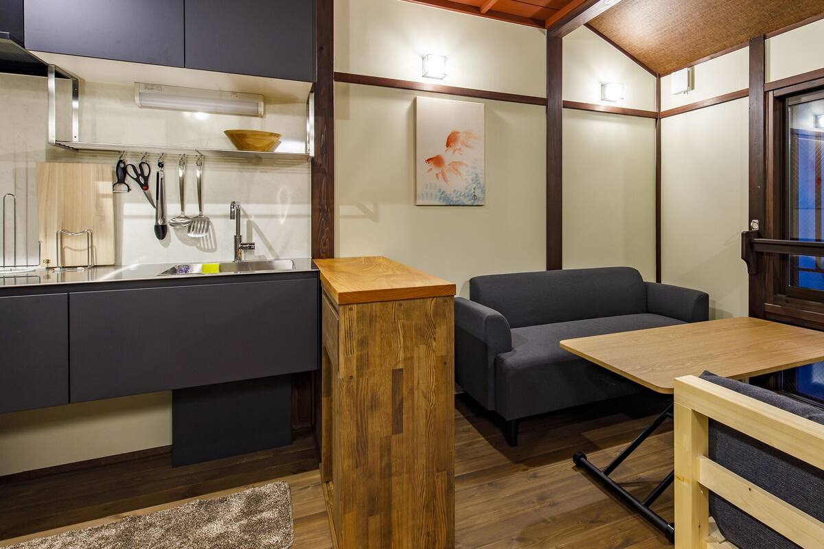 A compact kitchen with modern appliances, a dining area with a wooden table, and a sofa with a view through a large window.