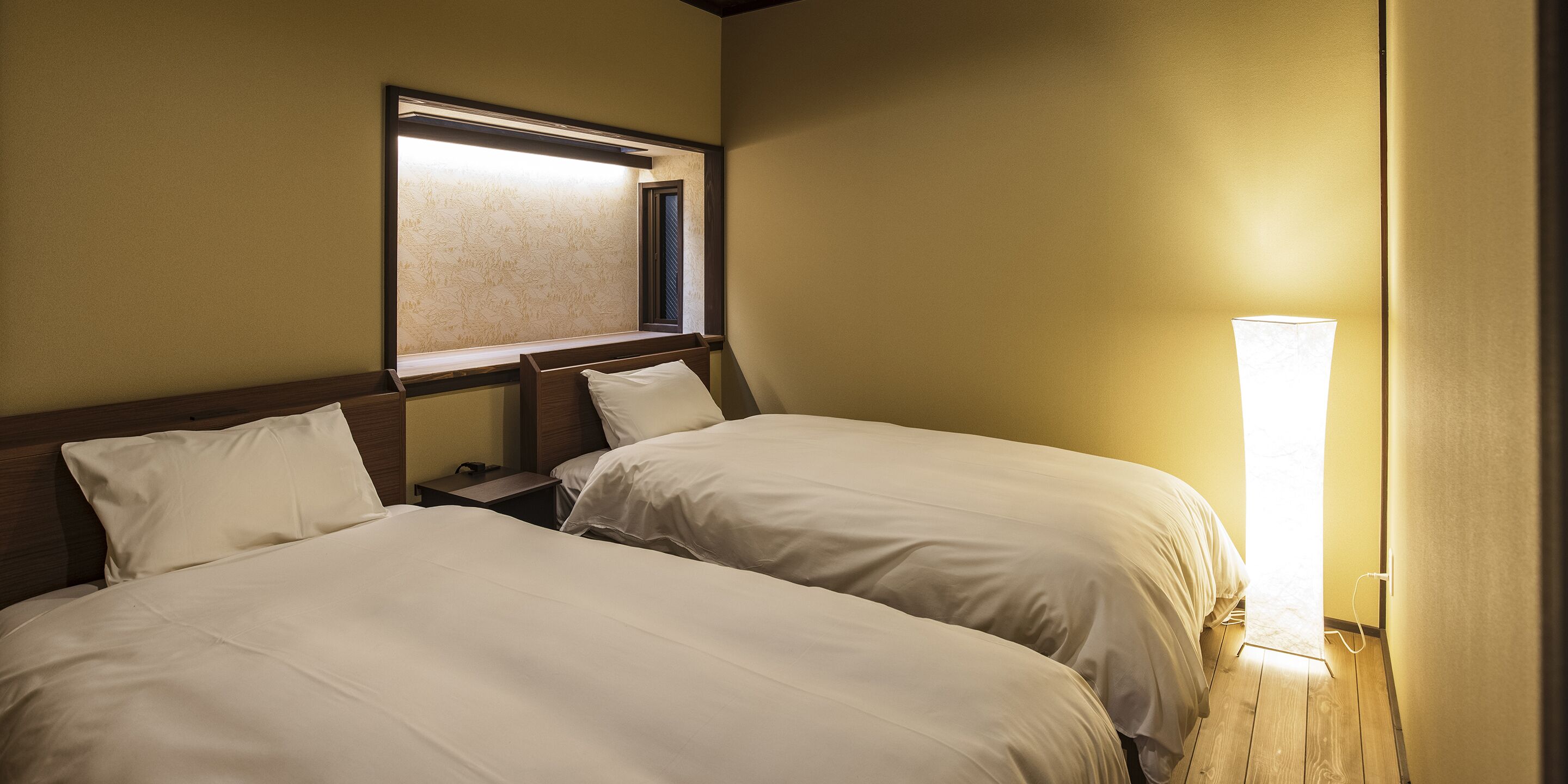 A hotel room with two beds, a nightstand, a lamp, and a window with blinds.