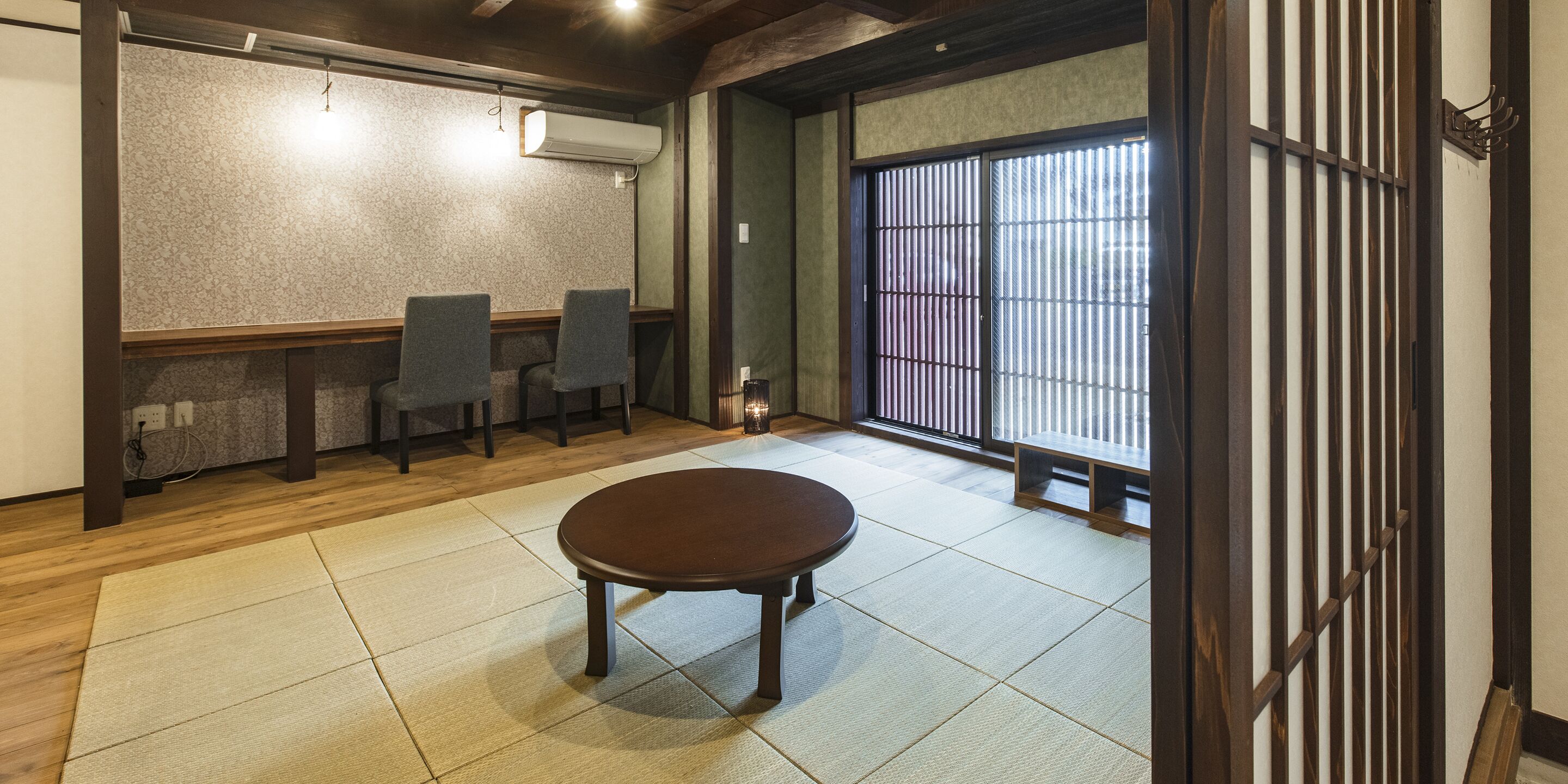 Una tradizionale stanza giapponese con pavimento in tatami, un tavolino in legno, due sedie, una porta scorrevole e una vista attraverso la porta.