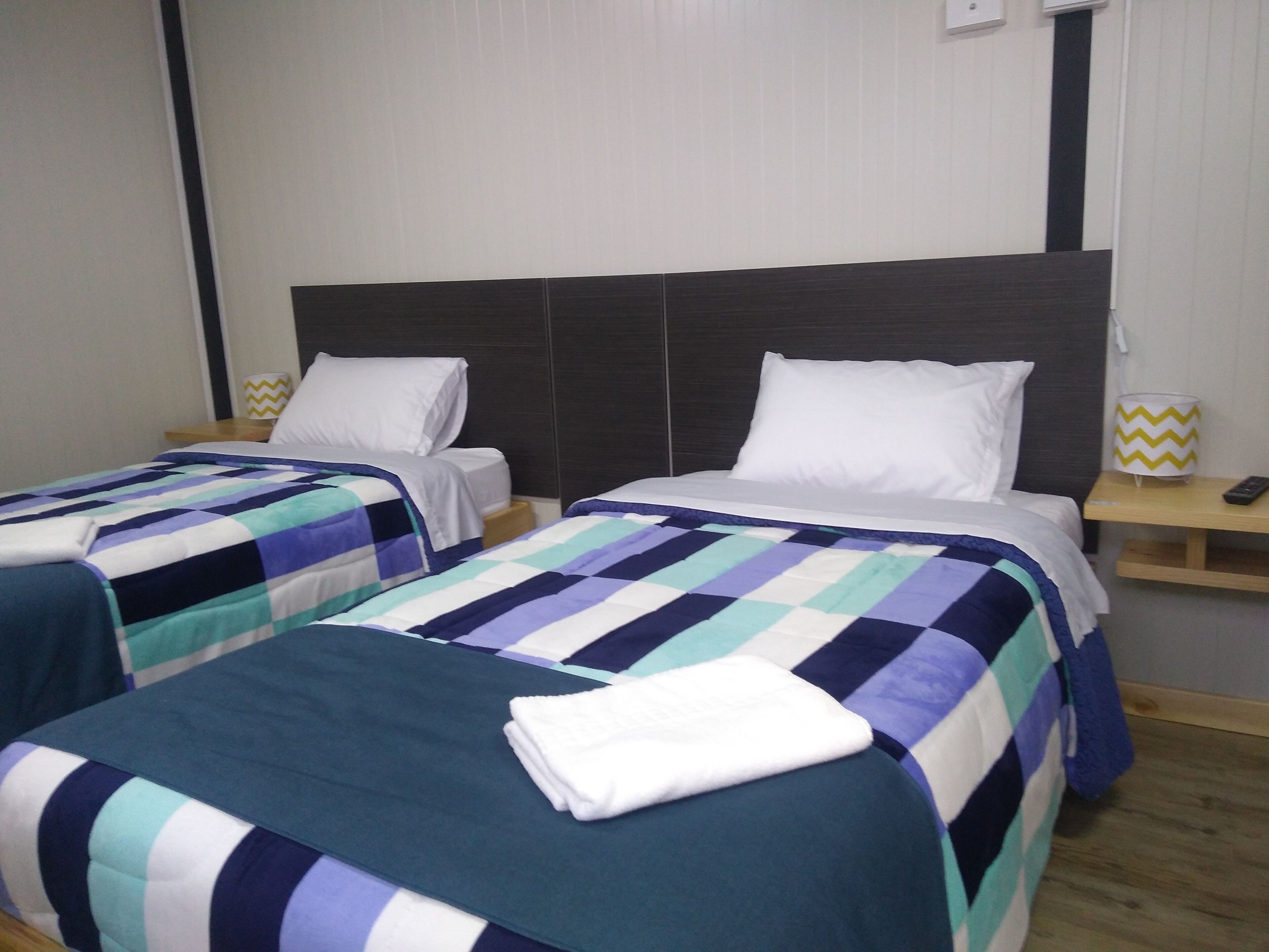 A hotel room with two beds, each with a checkered bedspread and white pillows. There are bedside tables with lamps and a wall-mounted light fixture.