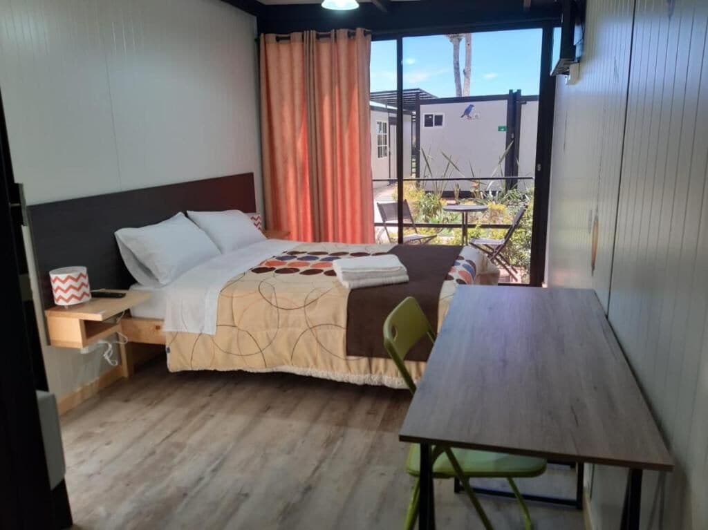 Une pièce comprenant un lit, une table et une chaise. Il y a une fenêtre avec des rideaux, ainsi qu’un balcon d’où l’on peut admirer les bâtiments.