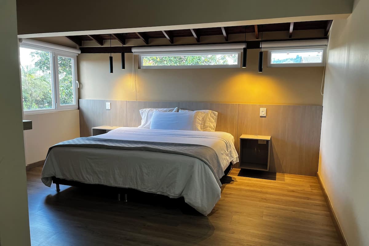 A modern bedroom with a large bed, wooden headboard, bedside tables, and a view of greenery through a window.