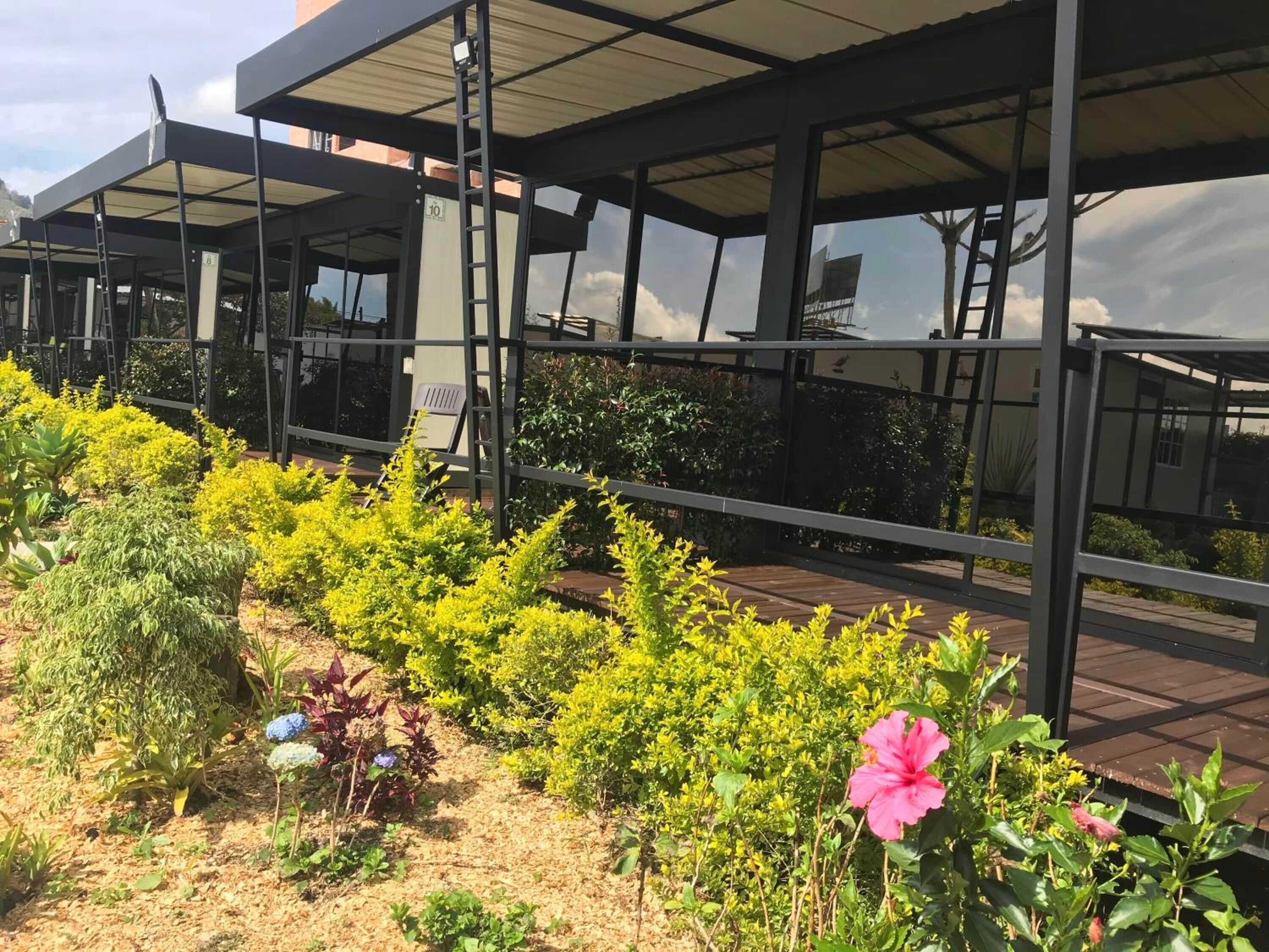 A covered outdoor area with a metal structure, numbered signs, and a variety of plants including yellow flowers and a pink hibiscus.