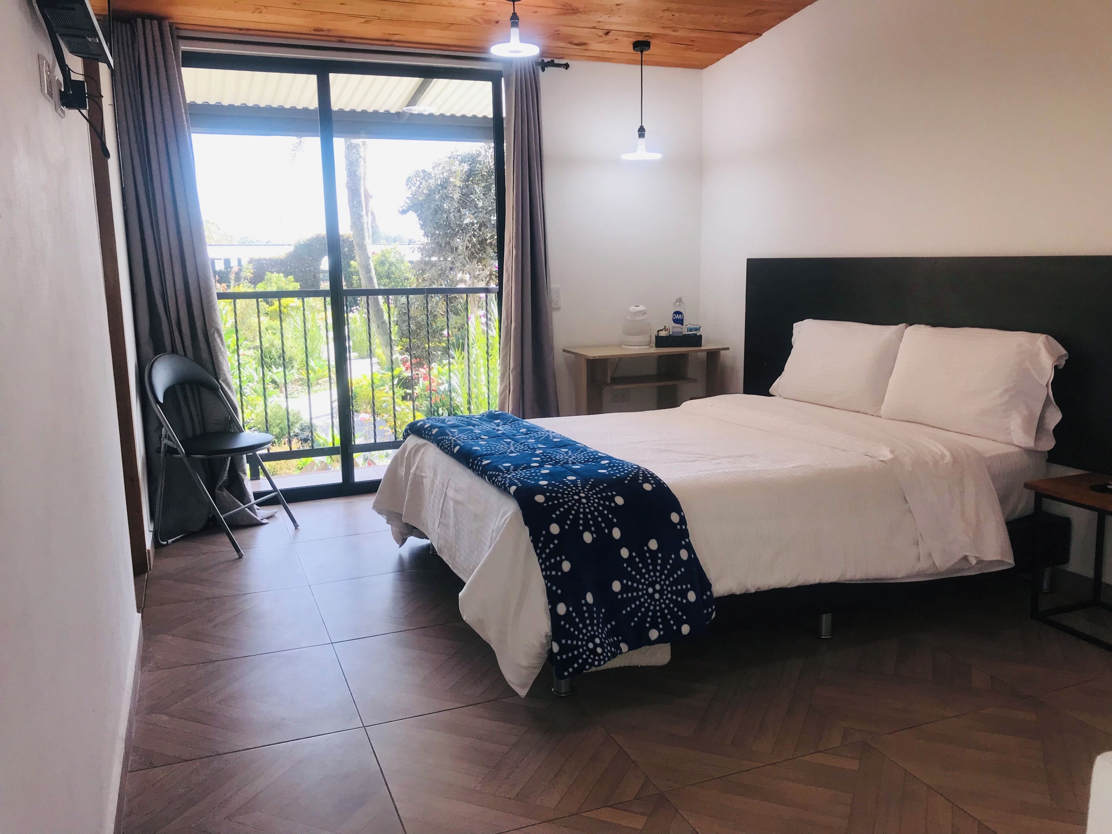 A bedroom with a bed, a chair, a nightstand, a lamp, and a view of greenery outside.