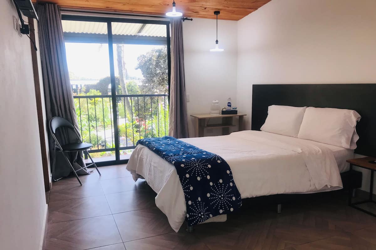 A bedroom with a bed, a chair, a nightstand, a lamp, and a view of greenery outside.