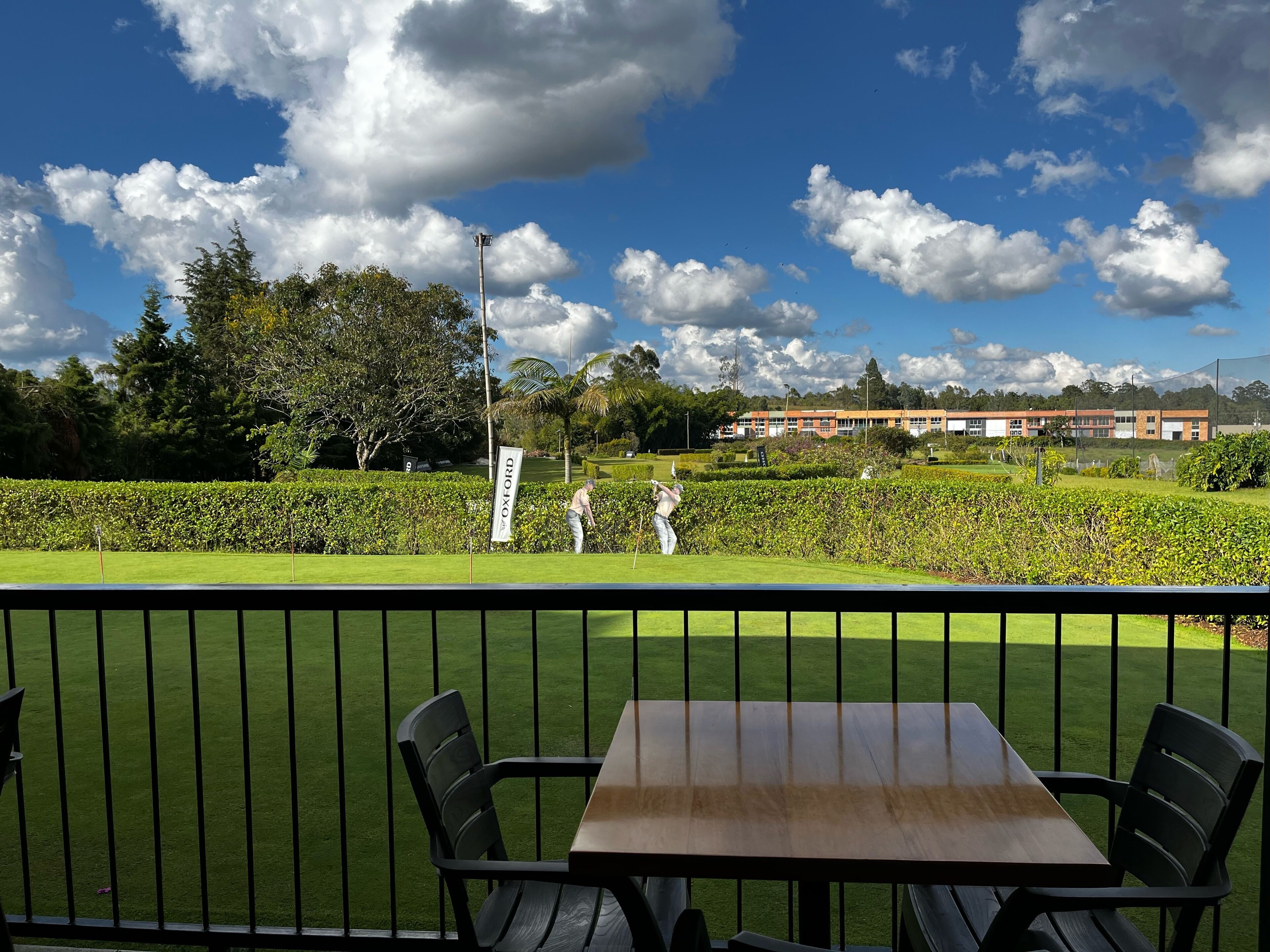Un campo da golf con un tavolo e delle sedie in primo piano, un cartello con la scritta 'OXFORD' sullo sfondo e un edificio con diverse finestre.