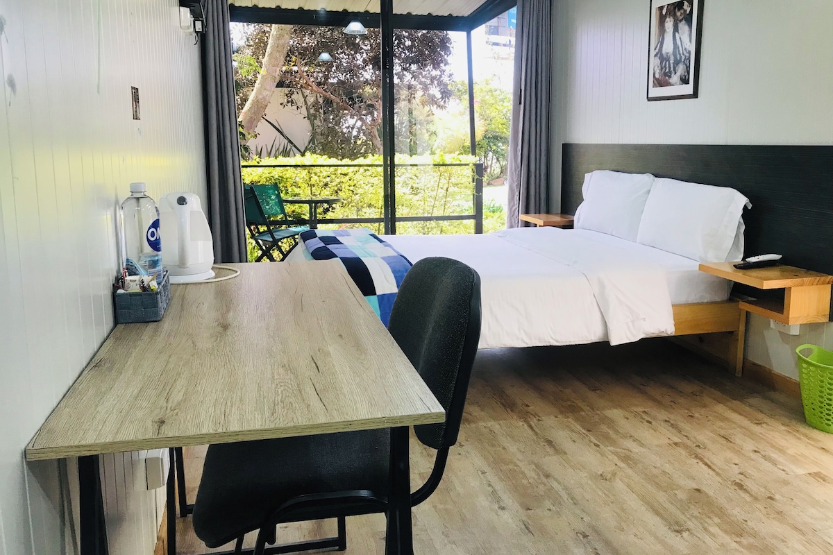 A hotel room with a bed, a desk, a chair, a nightstand, a window with curtains, and a view of greenery outside.