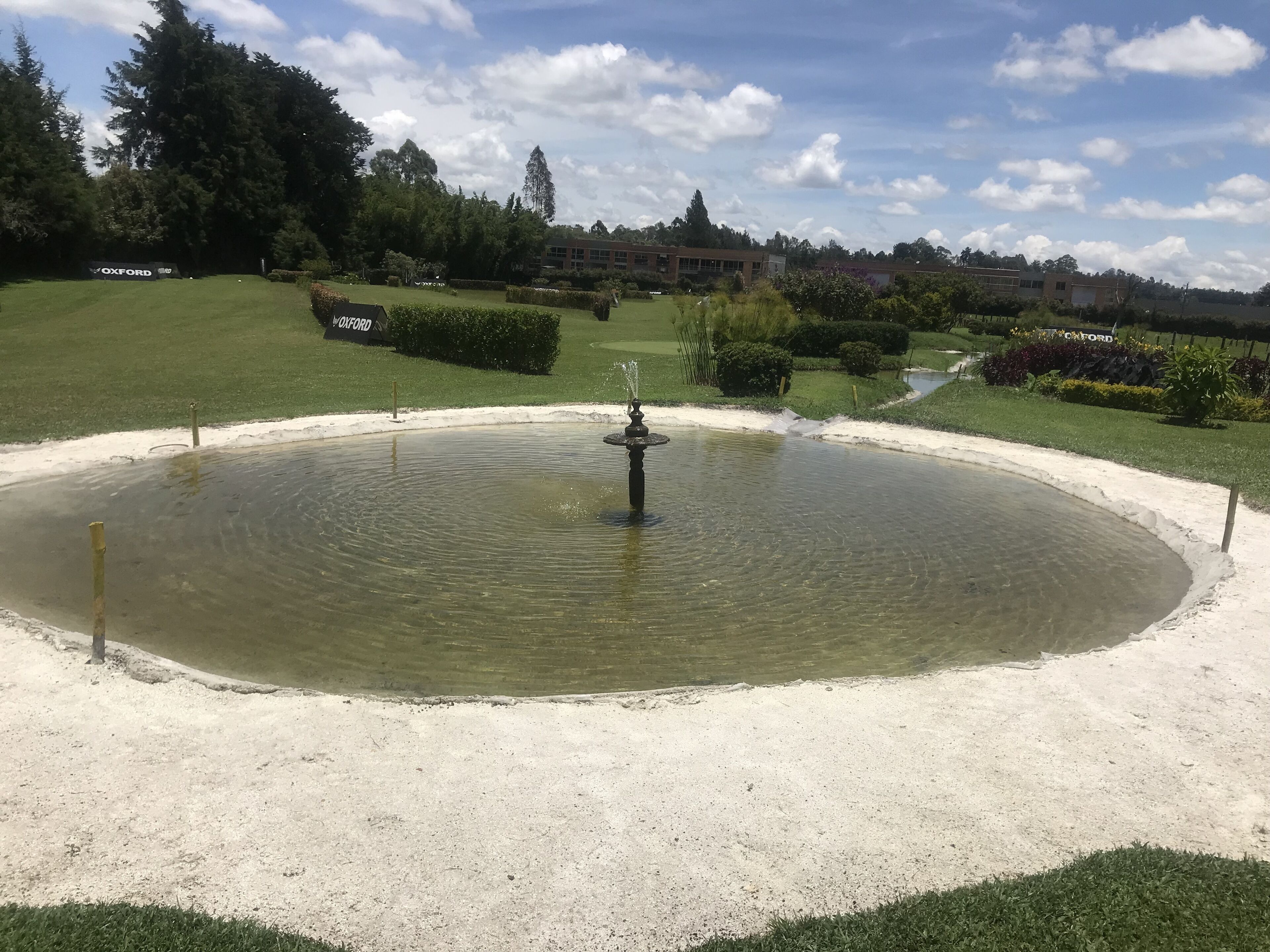 A circular water feature with a fountain in the center, surrounded by a sandy area and greenery.
